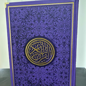May include: A purple hardcover book with a gold border and intricate black floral design. The cover features a gold circle with Arabic calligraphy in the center.