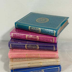 May include: A stack of six colorful hardcover books with gold accents and Arabic text on the covers. The books are arranged in a descending order of size, with the largest book on top and the smallest book on the bottom. The colors of the books are teal, pink, purple, peach, beige, and blue.