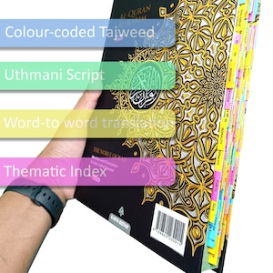 May include: A black and gold Quran with color-coded Tajweed, Uthmani script, and word-to-word translation. The book features a thematic index and is held open to show the colorful tabbed pages. The cover has gold floral patterns.