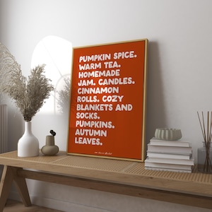 Könnte beinhalten: Ein orangefarbener gerahmter Druck mit weißem Text, der herbstliche Artikel auflistet: Kürbisgewürz, warmer Tee, hausgemachte Marmelade, Kerzen, Zimtschnecken, gemütliche Decken und Socken, Kürbisse und Herbstblätter. Der Druck wird auf einem Holztisch angezeigt.