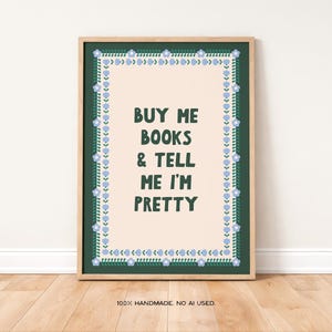 Könnte beinhalten: Ein gerahmter Kunstdruck mit dem Text "BUY ME BOOKS & TELL ME I'M PRETTY" in Dunkelgrün. Der Druck hat einen hellpfirsichfarbenen Hintergrund mit einem dunkelgrünen Rand, der mit blauen floralen Akzenten verziert ist. Der Rahmen ist hellholzfarben.