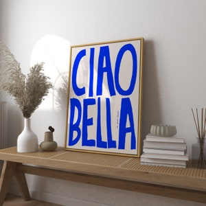 May include: A framed art print with the words "CIAO BELLA" in bold blue letters on a cream background. The artwork is in a gold frame and rests on a wooden bench with other decorative items.