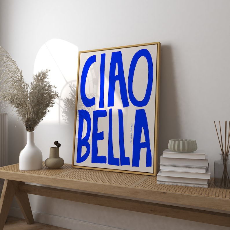 May include: A framed art print with the words "CIAO BELLA" in bold blue letters on a cream background. The artwork is in a gold frame and rests on a wooden bench with other decorative items.