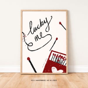 Pode incluir: Impressão de arte emoldurada com fundo creme. A obra apresenta as palavras "lucky me" em escrita preta, juntamente com fósforos espalhados e uma caixa de fósforos vermelha com um coração. A moldura é de cor de madeira clara.