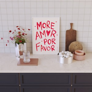 May include: A white framed print with the text "More Amor Por Favor" in pink lettering with a red heart. The print is on a white tiled countertop with a vase of pink flowers, a glass, a book, a bowl of white stones, and a wooden chopping board.