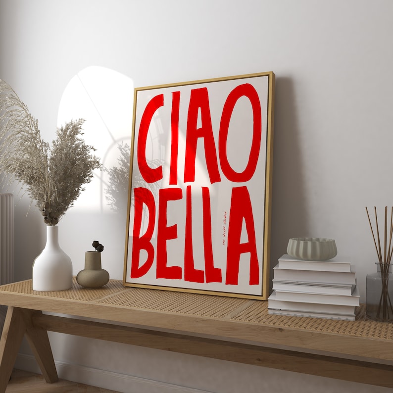 May include: A framed art print with the words "CIAO BELLA" in bold red letters on a cream background. The artwork is in a gold frame and is displayed on a wooden bench with decorative items.