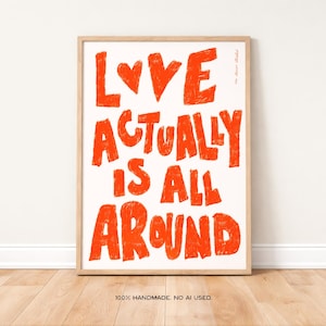 May include: A framed art print with the words "LOVE ACTUALLY IS ALL AROUND" in bright orange, handwritten-style lettering. The artwork features a heart symbol above the word "LOVE" and is set against a white background. The frame is a light wood colour.