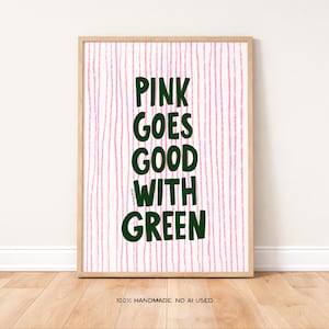 May include: A framed art print with the text "PINK GOES GOOD WITH GREEN" in dark green letters. The background has vertical pink lines. The frame is a light wood color. The bottom of the print reads "100% HANDMADE. NO AI USED."
