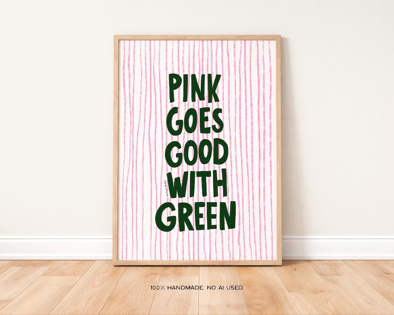 May include: A framed art print with the text "PINK GOES GOOD WITH GREEN" in dark green letters. The background has vertical pink lines. The frame is a light wood color. The bottom of the print reads "100% HANDMADE. NO AI USED."