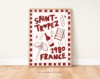 May include: A framed art print with a red and white design. The print features the words "SAINT-TROPEZ 1980 FRANCE" in a bold, handwritten style, along with illustrations of a wine bottle, beach umbrella, and lounge chair. The frame is a light wood color.