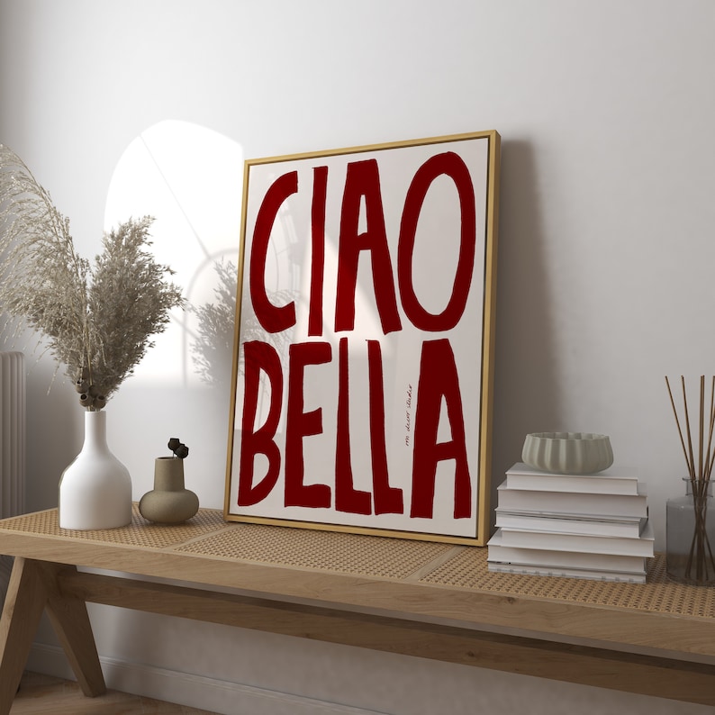 May include: A framed art print with the words "CIAO BELLA" in bold, dark red letters on a cream background. The artwork is in a gold frame and rests on a light wood bench with decorative items.