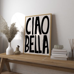 May include: A framed art print with the words "CIAO BELLA" in bold black letters on a cream background. The artwork is in a gold frame and rests on a wooden bench with decorative items.