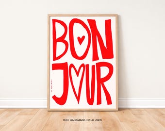May include: A framed art print with the word "BONJOUR" in bold red letters on a white background. The "O" in "BONJOUR" is replaced with a heart symbol. The print is in a light wood frame and is displayed against a white wall.