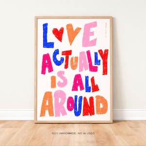 May include: A framed art print with the words "LOVE ACTUALLY IS ALL AROUND" in colorful, bold letters. The artwork features a heart and the text is in shades of blue, orange, pink, and red. The frame is a light wood color.