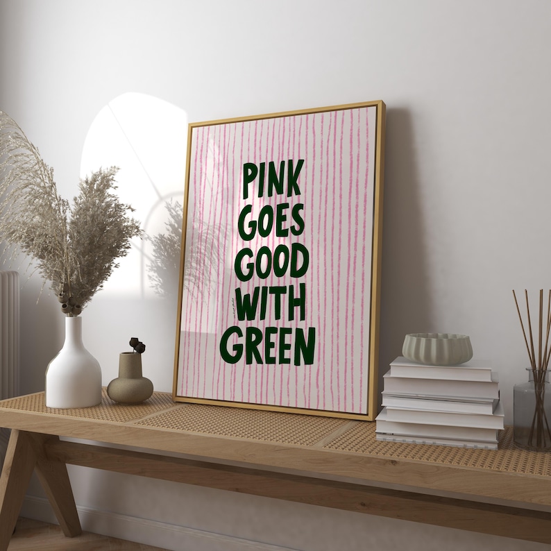 May include: A framed art print with the text "PINK GOES GOOD WITH GREEN" in dark green letters. The background has vertical pink stripes. The frame is gold. The print is displayed on a wooden bench.