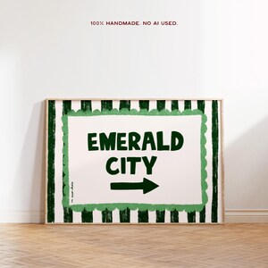 以下が含まれることがあります： 白い背景に緑色の「EMERALD CITY」の文字と右向きの矢印が描かれた額入りのアートプリント。フレームは緑と白のストライプ模様。フレーム上部には「100% HANDMADE. NO AI USED.」の文字。