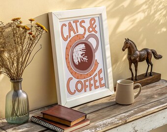 May include: A framed print with the words "CATS & COFFEE" in orange lettering. The artwork features a cat-shaped coffee cup design. A brown mug, books, a vase with dried flowers, and a horse statue are on a wooden surface.