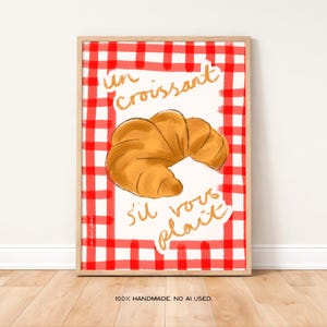 Op de afbeelding: Een ingelijste print met een gouden croissant illustratie op een rood-wit geblokte achtergrond. Gouden letters lezen "un croissant s'il vous pla&icirc;t". De zin "100% HANDMADE. NO AI USED." staat onderaan.