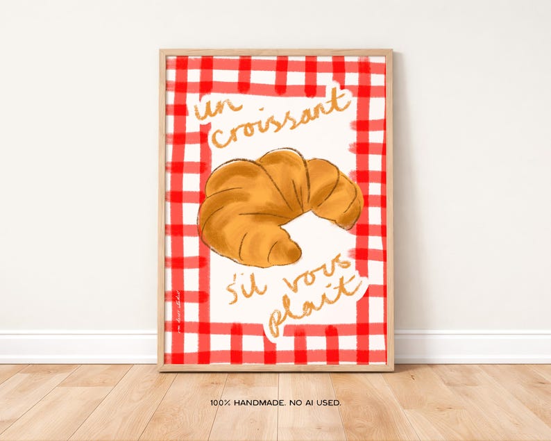 Op de afbeelding: Een ingelijste print met een gouden croissant illustratie op een rood-wit geblokte achtergrond. Gouden letters lezen "un croissant s'il vous pla&icirc;t". De zin "100% HANDMADE. NO AI USED." staat onderaan.