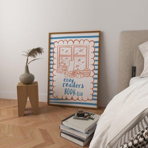 May include: A framed poster with a cosy illustration. The artwork features a window scene with a cat, books, and pumpkins, with the text "cozy readers BOOK CLUB" in blue and red. The frame is brown, and the background has blue and white stripes.