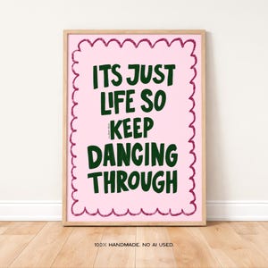 May include: A framed art print with a pink background and dark green text that reads "ITS JUST LIFE SO KEEP DANCING THROUGH". The print has a decorative pink border and is in a light wood frame. The bottom of the print reads "100% HANDMADE. NO AI USED."