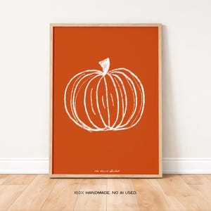 May include: A framed art print featuring a white sketched pumpkin on an orange background. The artwork is in a wooden frame and is displayed against a white wall and wooden floor. The text "100% HANDMADE. NO AI USED." is at the bottom.