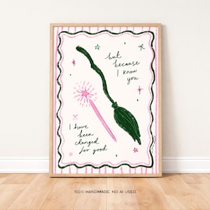 May include: A framed art print with a whimsical illustration. The artwork features a green broom and a pink wand with a starburst. Text reads "but because I knew you, I have been changed for good." The print is framed in a light wood frame.