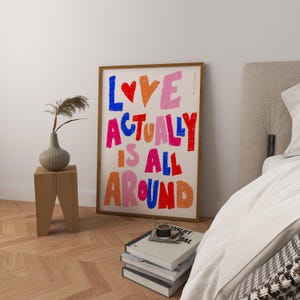 May include: A framed art print with the text "LOVE ACTUALLY IS ALL AROUND" in colourful, hand-drawn letters. The letters are in shades of pink, red, blue, and orange. The print is hanging on a white wall in a bedroom setting.