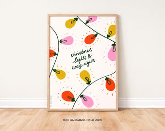 May include: A framed print featuring a whimsical illustration of Christmas lights in yellow, pink, and orange against a cream background. The text "christmas lights & cosy nights" is written in green cursive. The frame is a light wood color.