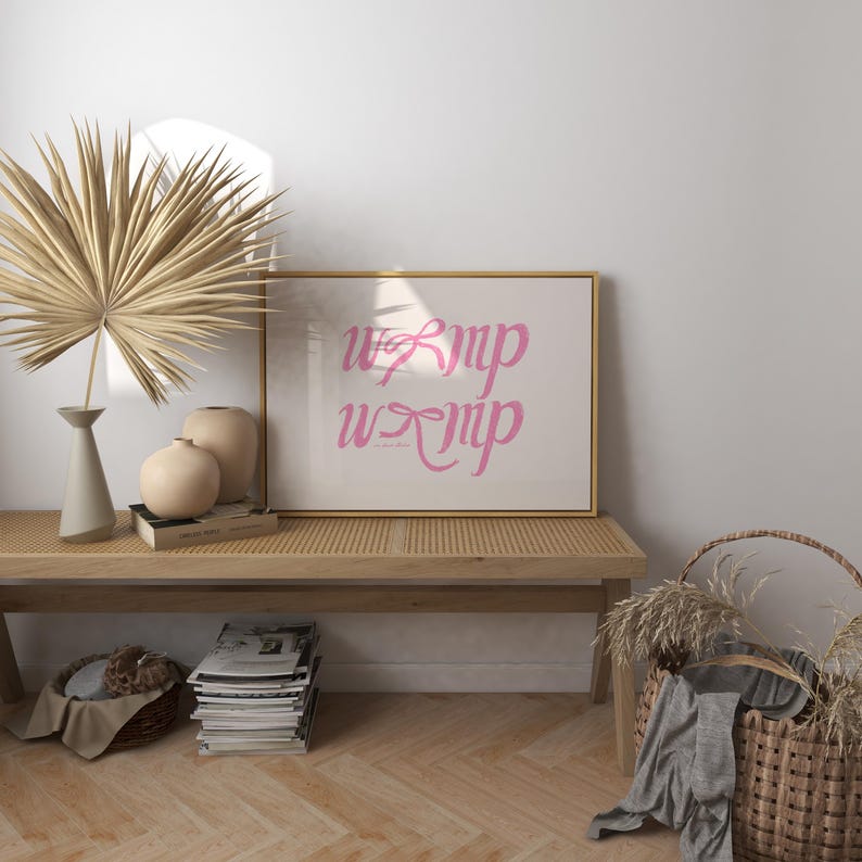 May include: A framed print with the word "WIMP" written twice in pink cursive letters on a white background. The frame is gold and the print is sitting on a wooden bench with a woven rattan basket and dried grass.