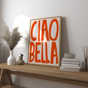May include: A framed art print with the words "CIAO BELLA" in bold orange letters on a white background. The artwork is in a gold frame and rests on a wooden bench with other decorative items.