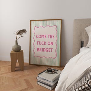 May include: A framed art print with the text "COME THE FUCK ON BRIDGET" in pink, set against a white background with green vertical stripes. The print is in a wooden frame and is displayed in a bedroom setting.