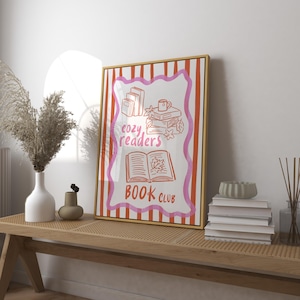 May include: A framed print with a gold border featuring the words "cozy readers book club." The artwork includes illustrations of books, a mug, and autumn leaves. The background has pink and orange stripes.