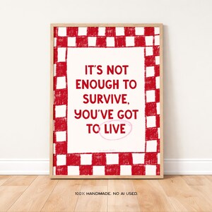 May include: A framed print with a red and white chequered border. The text "IT'S NOT ENOUGH TO SURVIVE, YOU'VE GOT TO LIVE" is centered on a cream background. The words "100% HANDMADE. NO AI USED" are at the bottom.