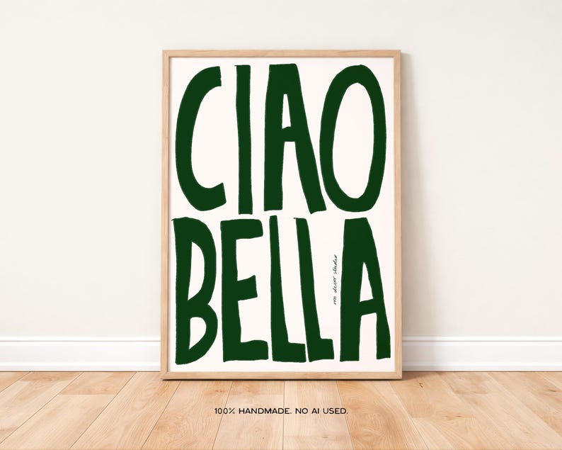 May include: A framed art print with the words "CIAO BELLA" in bold, dark green letters on an off-white background. The print is in a light wood frame and is displayed against a white wall and wooden floor.