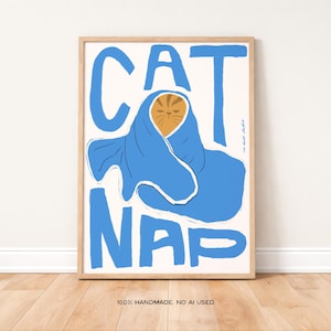 Könnte beinhalten: Gerahmter Kunstdruck mit einer Illustration einer Katze in einer blauen Decke. Die Worte "CAT NAP" stehen in großen, blauen Buchstaben über und unter der Katze. Das Kunstwerk ist vor einem weißen Hintergrund.