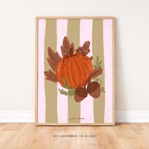 May include: A framed art print depicting an orange pumpkin with brown leaves and acorns, set against a backdrop of vertical olive green and pink stripes. The artwork is displayed on a wooden floor.