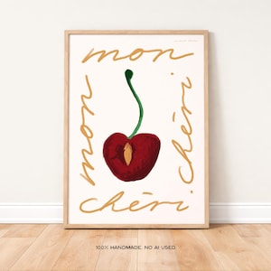 May include: A framed art print featuring a stylized cherry illustration. The cherry is a deep red with a green stem, set against a cream background. Gold cursive text reads "mon chéri" around the cherry. The frame is a light wood color.
