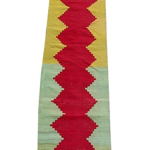 May include: A long, narrow rug with a geometric pattern. The rug is made of wool and has a red, yellow, and green colour scheme.