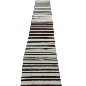 Puede incluir: Una alfombra larga y estrecha con fondo blanco y rayas negras. La alfombra tiene algunas rayas rojas y marrones cerca de la parte superior.