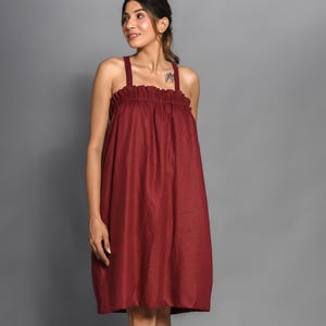 May include: A burgundy-colored dress with a gathered neckline and wide shoulder straps. The knee-length dress has a loose, flowing silhouette. The model is wearing clear sandals. The background is a solid gray.