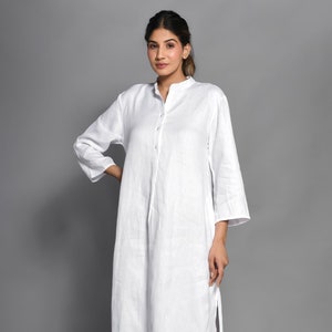 Könnte beinhalten: Ein weißes Leinenkleid mit Kragen, langen Ärmeln und Knopfleiste. Das Kleid hat einen seitlichen Schlitz und eine lockere Passform.