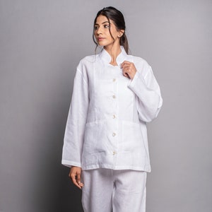 Op de afbeelding: Een witte linnen pyjamaset met een getailleerd topje en lange broek. Het topje heeft een knoopsluiting aan de voorkant en een klepzak aan de linkerkant.