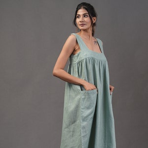 Può includere: Abito midi verde chiaro senza maniche, con scollo quadrato e due ampie tasche frontali. L'abito ha una silhouette ampia e fluida ed è realizzato in un tessuto morbido e strutturato. La modella indossa sandali trasparenti con tacco. L'abito è lungo circa 100 cm.