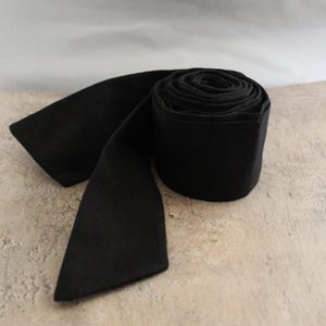 May include: A solid black fabric ribbon rolled into a tight cylinder with one end extending outwards. The material has a matte, uniform texture and a consistent deep black colour throughout the entire length of the coiled textile strip.