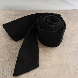 May include: A solid black fabric sash or belt rolled into a circular coil with one end extending outward. The material has a matte, uniform texture and a consistent deep black colour throughout the entire length of the long, flat textile strip.