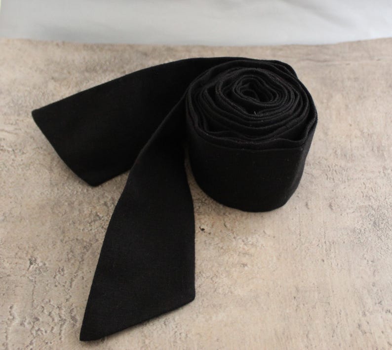May include: A solid black fabric sash or belt rolled into a circular coil with one end extending outward. The material has a matte, uniform texture and a consistent deep black colour throughout the entire length of the long, flat textile strip.