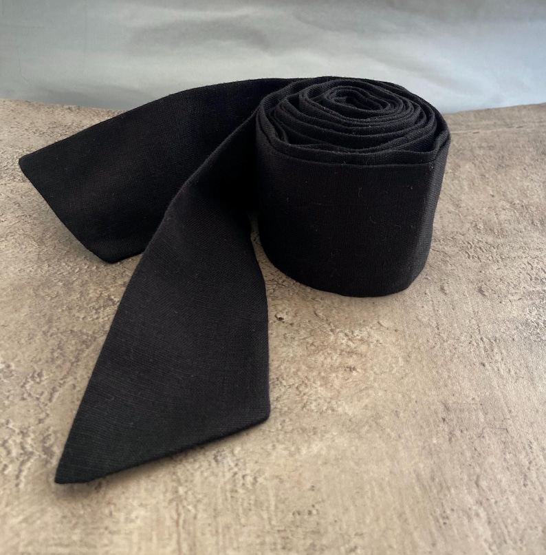 May include: A solid black fabric strip rolled into a cylindrical shape with one end extending outwards. The material has a matte finish and a consistent dark black colour throughout the entire length of the rolled textile band.