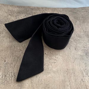 May include: A solid black fabric sash or ribbon rolled into a circular coil. The material has a matte texture and a consistent dark black colour throughout. One end of the long, flat strip of fabric extends outward from the side of the tight roll.