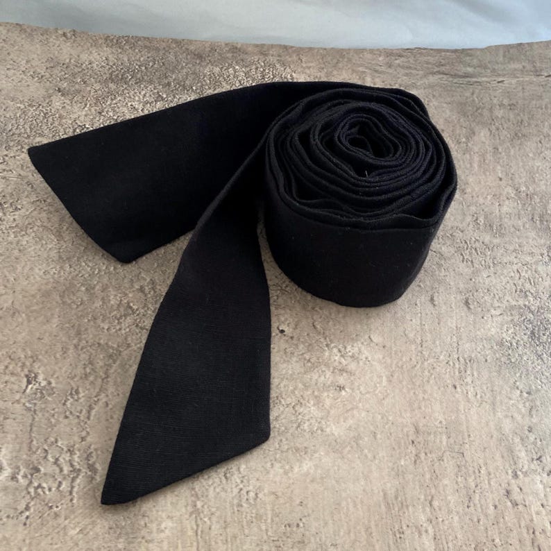 May include: A solid black fabric sash or ribbon rolled into a circular coil. The material has a matte texture and a consistent dark black colour throughout. One end of the long, flat strip of fabric extends outward from the side of the tight roll.